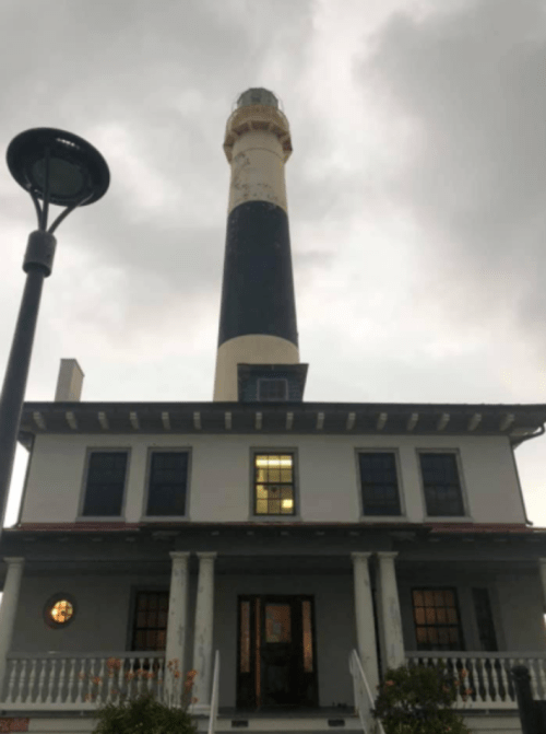 A tall lighthouse stands behind a historic building, under a cloudy sky, with a lamp post in the foreground.