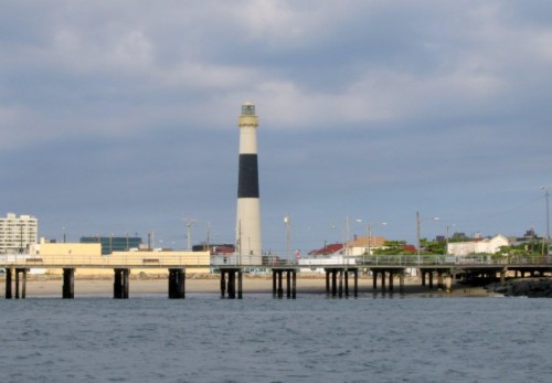 A lighthouse stands tall near a pier, with buildings in the background and calm waters in the foreground.