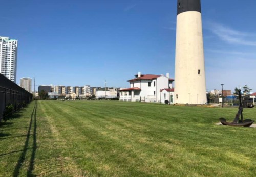 A grassy area with a lighthouse and a white building, surrounded by a city skyline under a clear blue sky.