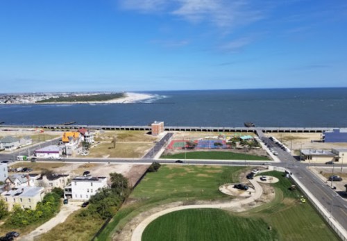 Aerial view of a coastal area with a beach, water, and a park featuring a playground and green spaces.