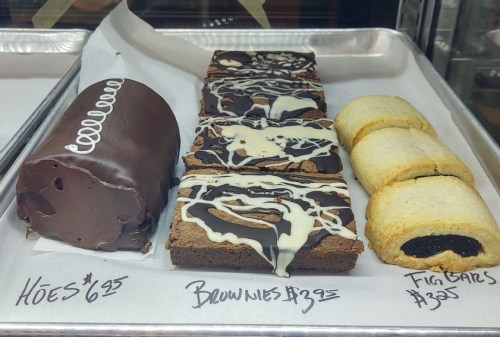 A display of desserts: chocolate rolls, brownies with white drizzle, and fig bars, all with prices listed.