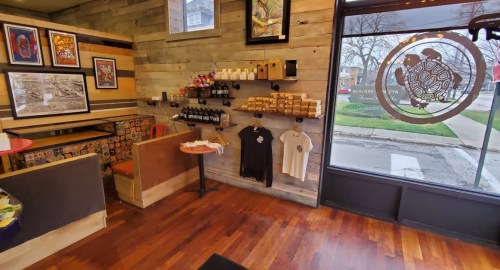 Cozy shop interior with wooden walls, art on display, and merchandise like shirts and bottles on shelves.