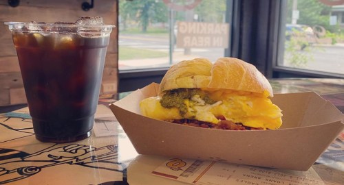 A sandwich with eggs and green sauce next to a cup of iced coffee on a table in a cafe.