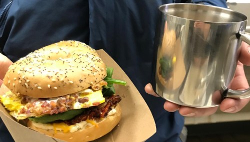 A person holds a sesame bagel sandwich with egg, cheese, and greens in one hand and a metal mug in the other.