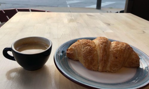 A cup of coffee next to a plate with a golden-brown croissant on a wooden table.