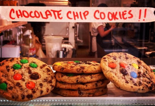 A display of large chocolate chip cookies with colorful candy pieces, labeled "Chocolate Chip Cookies !!" in the background.
