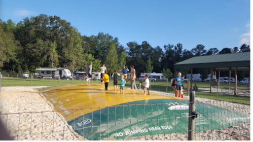Children and adults play on a large inflatable bounce pad at a park, surrounded by trees and RVs in the background.