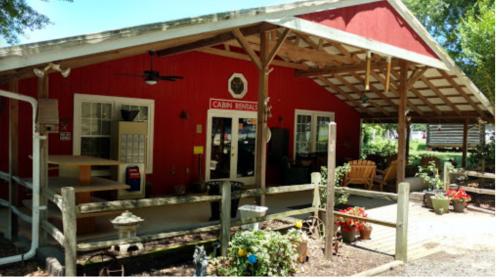 A red cabin rental building with a porch, surrounded by flowers and greenery, under a wooden roof.
