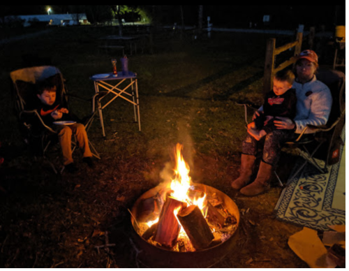 A cozy campfire scene with two children and an adult sitting in chairs, enjoying the warmth of the flames at night.