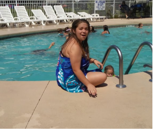 A girl in a blue swimsuit sits by a pool, smiling, while others swim in the background.