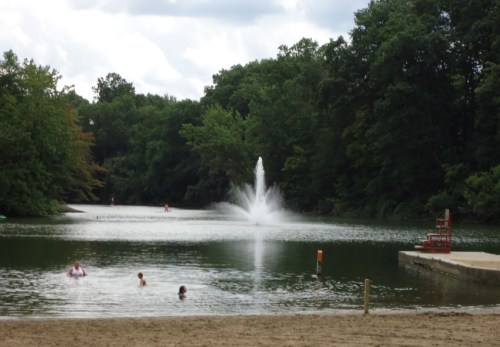 A serene lake with a fountain, surrounded by trees, and people swimming near the sandy shore.