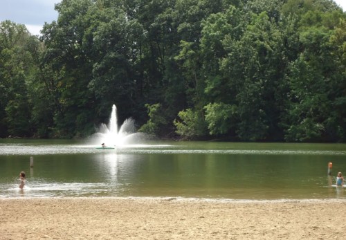 A serene lake scene with a fountain, surrounded by trees, and children playing in the sandy area by the water.