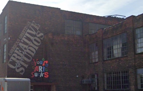 A brick building with large windows, featuring a colorful sign that reads "Your Art Sucks" and "Creative Studios."