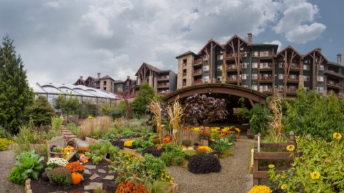 A vibrant garden with pumpkins and flowers, set against a backdrop of a large lodge under a cloudy sky.