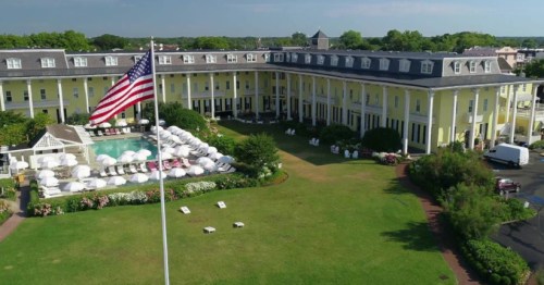 Aerial view of a yellow hotel with a pool and American flag in the foreground, surrounded by green lawns and trees.