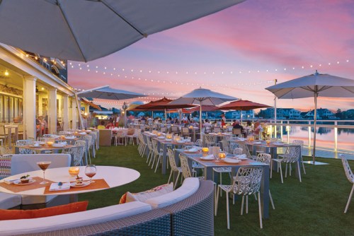 Outdoor dining area with tables, umbrellas, and string lights by a waterfront at sunset.
