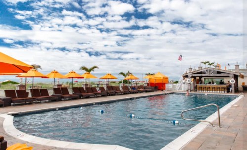 A sunny pool area with lounge chairs, orange umbrellas, and a bar, set against a cloudy sky and palm trees.
