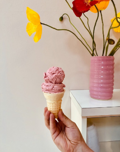 A hand holding a cone with two scoops of pink ice cream, next to a vase of colorful flowers.