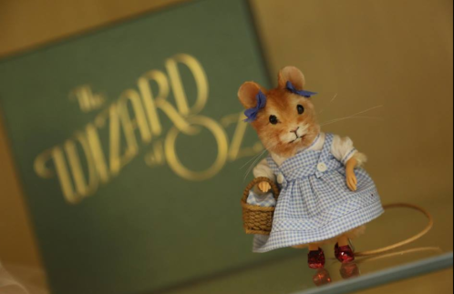 A cute mouse in a blue gingham dress and red shoes stands next to a "Wizard of Oz" book.