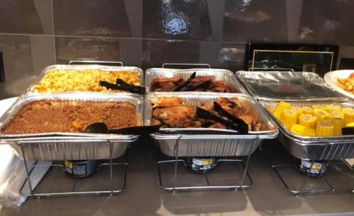 A buffet display with trays of macaroni and cheese, baked beans, roasted chicken, and corn on the cob.