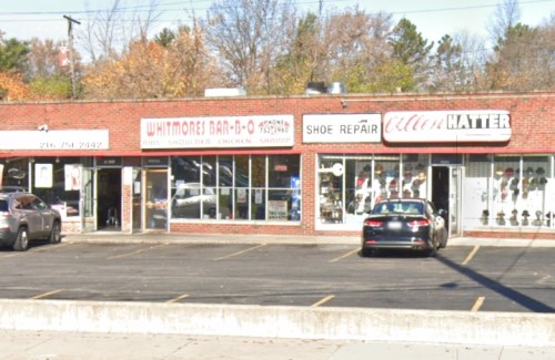 A storefront with "Whitmore's Bar-B-Q," "Shoe Repair," and "Cutter's Matter" signs, surrounded by trees in autumn.