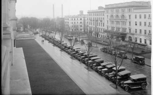 Historic black-and-white photo of a street lined with vintage cars, trees, and buildings on a rainy day.