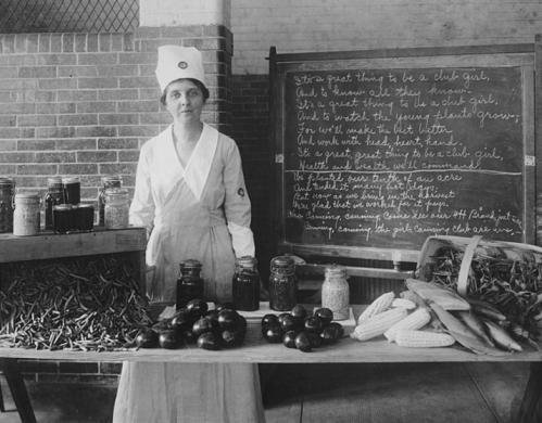 A woman in a chef's hat stands behind a table of fresh produce and jars, with a chalkboard displaying handwritten text.