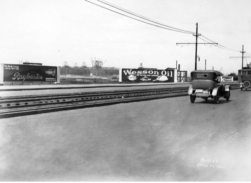 Black and white photo of a street with cars, railroad tracks, and vintage advertisements for Wesson Oil and Raybestos.