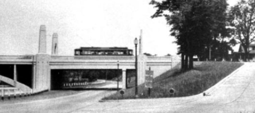 A vintage black-and-white photo of a bridge with a streetcar passing over it, surrounded by trees and a winding road.
