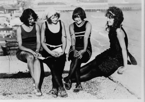 Four young women in vintage swimsuits sit together on a rooftop, with a beach and ocean in the background.