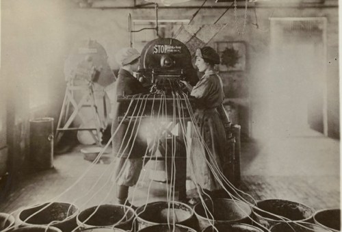 Two women operate a vintage machine with multiple wires in a factory setting, surrounded by large containers.
