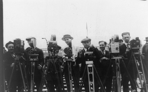 A group of men in suits standing behind vintage cameras on tripods, with a cloudy sky in the background.