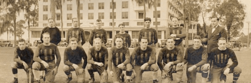 A vintage football team poses in front of a large building, wearing dark jerseys and standing on a grassy field.