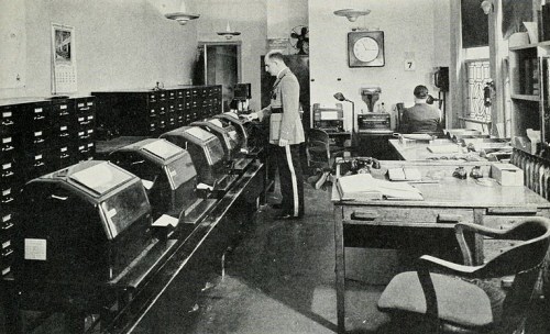 A vintage office with two men, one at a desk and another using a large machine, surrounded by filing cabinets.