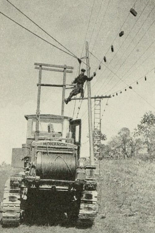 A worker climbs a utility pole using a ladder, while a large tractor is positioned below, surrounded by power lines.
