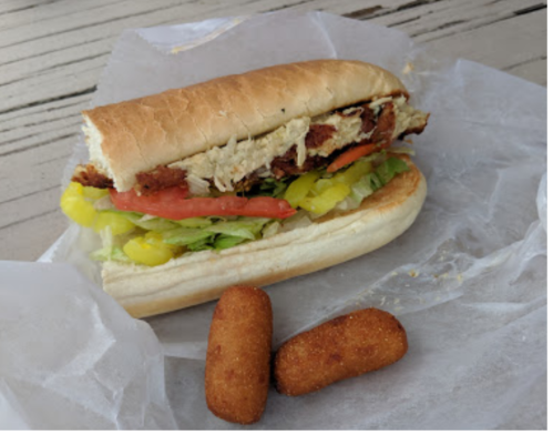 A sandwich with fried chicken, lettuce, tomato, and pickles, served with two golden croquettes on the side.