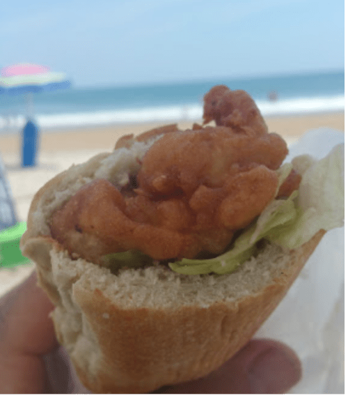 A close-up of a fried shrimp sandwich with lettuce, held against a beach backdrop with ocean waves.