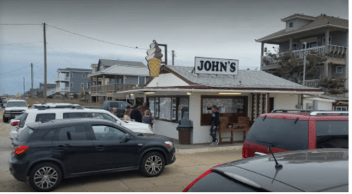 A busy ice cream shop named "John's" with cars parked outside and people waiting in line. Coastal buildings in the background.