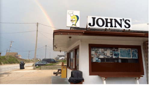 A restaurant named "John's" with a cartoon fish sign, under a rainbow, near a road and power lines.