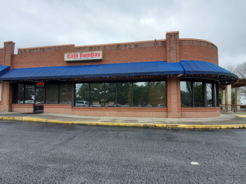 Exterior of Cafe Bombay featuring a blue awning, large windows, and an "OPEN" sign on a cloudy day.