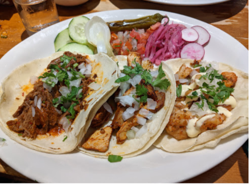 Three tacos on a plate, filled with meat, onions, cilantro, and served with cucumber, radishes, and pickled vegetables.