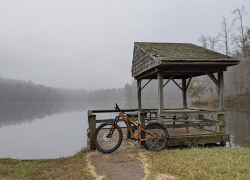 A mountain bike rests beside a wooden gazebo by a foggy lake, surrounded by trees and calm water.