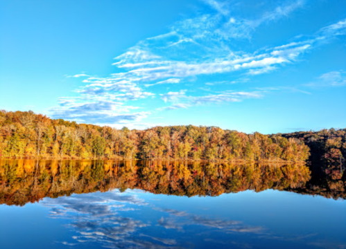 A serene lake reflects colorful autumn trees under a clear blue sky with wispy clouds.