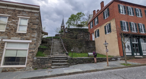 A stone building and a red brick building flank a set of stairs leading to a grassy area with a church steeple in the background.