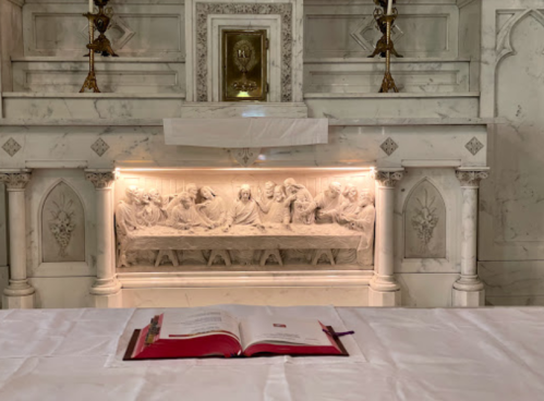 A marble altar with a relief of the Last Supper and an open book on a white tablecloth in the foreground.