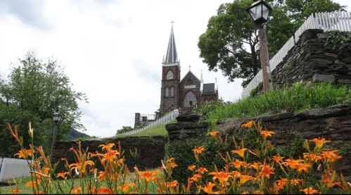 A church with a tall steeple stands behind vibrant orange flowers and green grass under a cloudy sky.