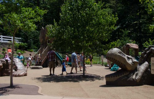 A playground with dinosaur-themed structures, children playing, and trees in a sunny outdoor setting.