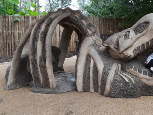 A large, artistic sculpture resembling a dinosaur skeleton, set in a natural outdoor environment.