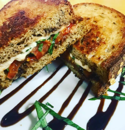 A close-up of a toasted sandwich with layers of tomato, greens, and sauce, served on a white plate with drizzled balsamic.