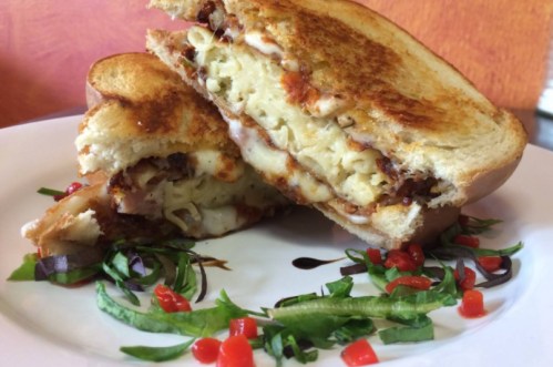 A grilled sandwich filled with melted cheese and pasta, served with fresh greens and diced red peppers on the side.